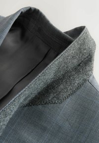 Blazer à carreaux gris avec une doublure intérieure de col douce et texturée et une doublure intérieure en tissu noir lisse, vu de dessus sur un fond blanc.