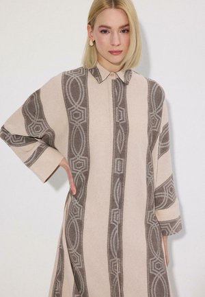 Blondynka ubrana w beżowo-szarą sukienkę z długim rękawem i kołnierzykiem, pozująca z jedną ręką na biodrze na jednolitym tle.