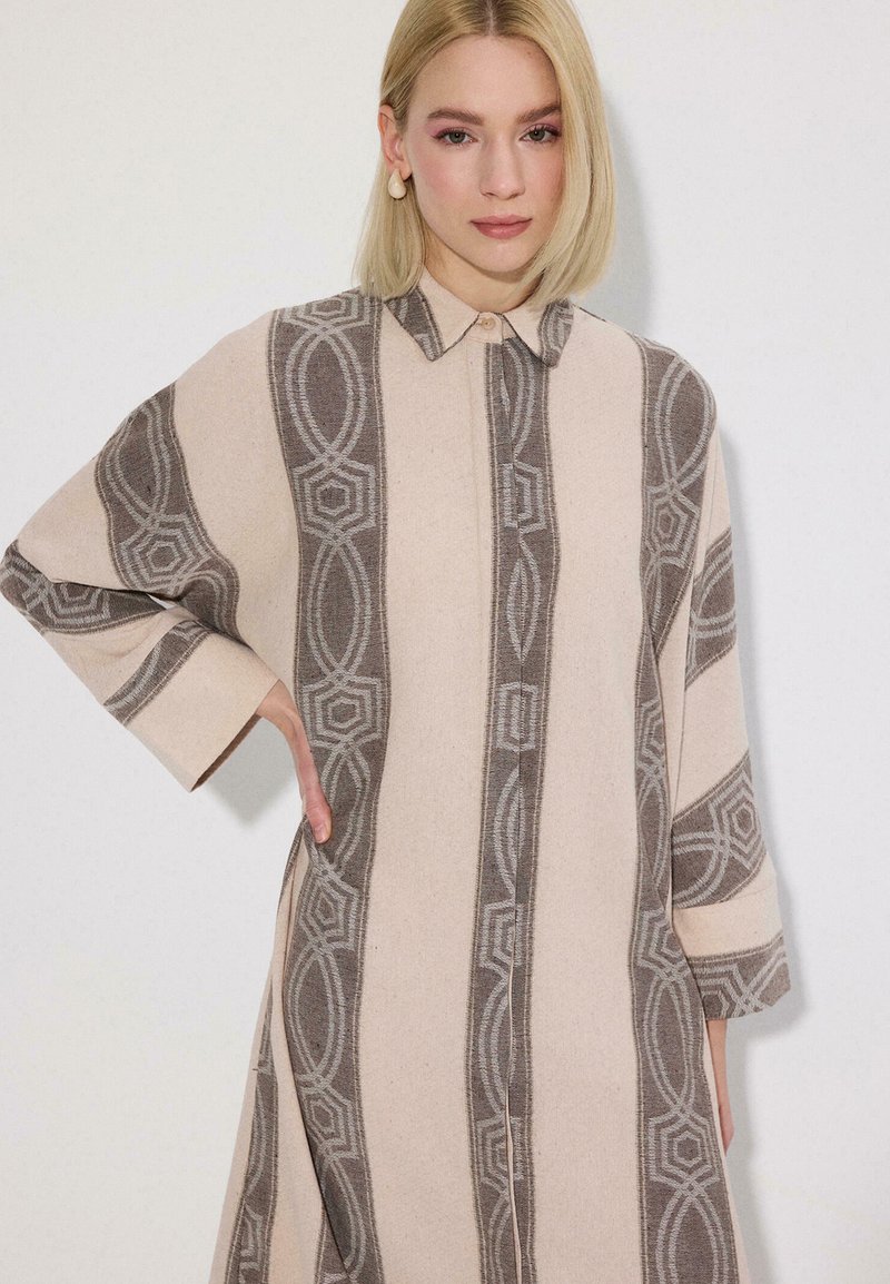 Femme blonde portant une robe à manches longues beige et grise à motifs avec un col, posant avec une main sur la hanche sur un fond uni.