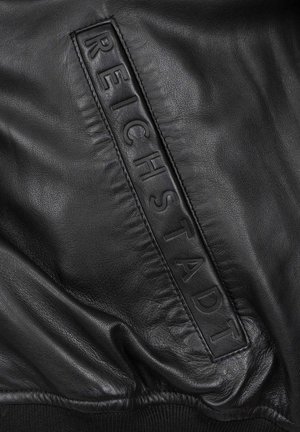 Veste en cuir noir avec un patch en relief bien en vue portant l'inscription "REICHSTADT", un tissu lisse et des détails en tissu froncé.