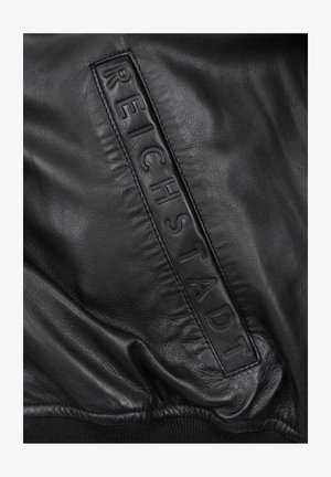 Schwarze Lederjacke mit einem auffälligen geprägten Patch, auf dem "REICHSTADT" steht, mit glatter Textur und gerafften Stoffdetails.