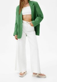 Groene linnen blazer over een wit cropped top, gecombineerd met wijdvallende witte jeans en beige sandalen met plateauzool. Details van getextureerde stof.
