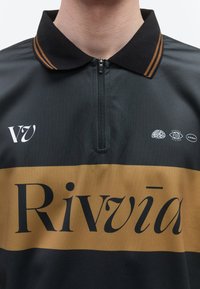 Μαύρο και καφέ πόλο πουκάμισο με φερμουάρ στη μέση, κείμενο «Rivvā» στο στήθος και μικρά εικονίδια εγκεφάλου, ματιού και κάψουλας πάνω από το κείμενο.