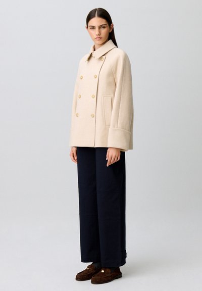 Donna con cappotto doppiopetto beige, pantaloni larghi scuri e mocassini marroni, in piedi davanti a uno sfondo semplice grigio chiaro.