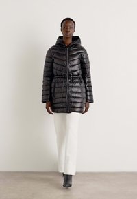 Doudoune noire avec des coutures horizontales, taille à cordon de serrage et zip avant, associée à un pantalon blanc et des bottes grises.