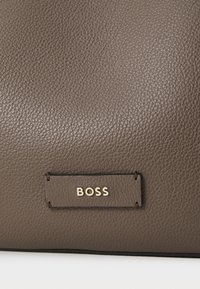 Sötétbarna bőr kézitáska, texturált felülettel. Arany "BOSS" logó található egy téglalap alakú címkén. Simább élek és strukturált forma jellemzi.