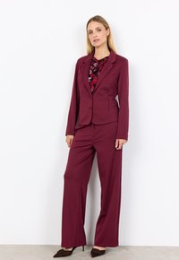 Frau in einem burgunderfarbenen Hosenanzug mit einem taillierten Blazer und weitem Hosenbein, kombiniert mit einem gemusterten rot-schwarzen Oberteil und dunklen Absätzen.