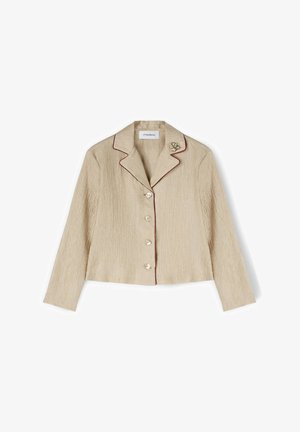 Blazer beige texturé à manches longues avec quatre boutons, bordure marron foncé sur le col et le devant, et une broche décorative sur le revers gauche.