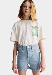 Ljusbeige bomulls-t-shirt med grönt grafiskt "RODEO"-motiv och blå jeansshorts. Shortsen har fickor fram och bak samt hög midja.