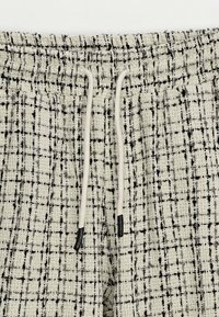 Pantalons en tissu texturé de couleur claire avec un motif à carreaux noirs, dotés d'une taille élastique et de cordons de serrage avec des embouts noirs.