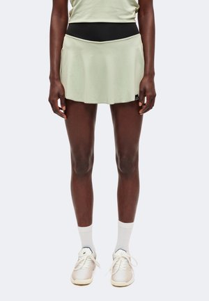 Personne portant une jupe de sport vert clair avec une ceinture noire, des chaussettes blanches montantes et des baskets blanches à lacets, debout, le dos droit.