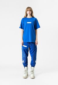Camiseta de algodón azul con un panel frontal y corte relajado, combinada con pantalones de chándal azules que presentan bordados de plumas y detalles en blanco. Botas blancas.