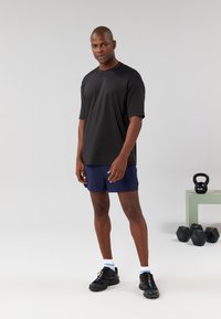 Črno kratkoročno majico in modre kratke hlače, ujemajoče se s črnimi supergami in belimi nogavicami. V ozadju je fitnes oprema.