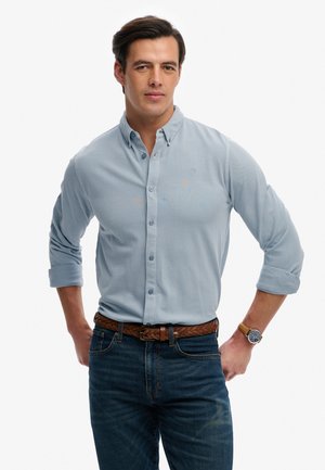 Mężczyzna w jasnozielonej koszuli z długim rękawem podwiniętym do łokci, plecionym brązowym paskiem, ciemnych dżinsach i zegarku na nadgarstku, stoi z rękami na biodrach.