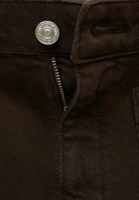 Brune denimbukser med en sølvmetal knap og lynlås. Stoffet har en glat tekstur med subtile syninger.