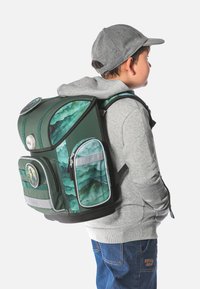 Zaino verde con motivo montano, spallacci regolabili, diverse tasche con zip e una toppa decorativa rotonda. Indossato da un bambino in una felpa grigia.