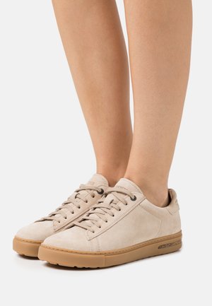 Beige suede sneakers with round toe, tonal laces, and tan rubber soles. Features a minimal design and subtle textured accents.
