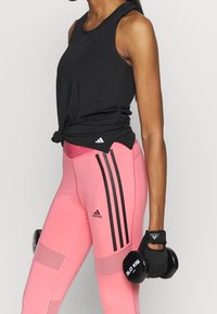 Czarna bezrękawnikowa koszulka sportowa z węzłowym detalem, zestawiona z różowymi legginsami z czarnymi paskami po bokach i akcentami z siateczki. Trzymając hantle.