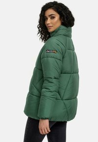 Groene pufferjack met ruitjespatroon, hoge kraag en elastische manchetten. Voorzien van een patch op de rechtermouw.