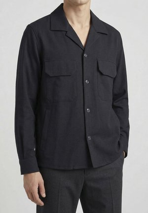 Man draagt een zwart overhemd met knopen, twee borstzakken en lange mouwen, gecombineerd met donkergrijze broek, neutrale achtergrond.