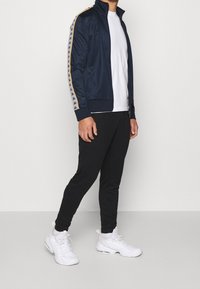 Marinblå zip-up jacka med kontrasterande guld- och vita ränder över axlarna, matchad med svarta avsmalnande byxor och vita sneakers.