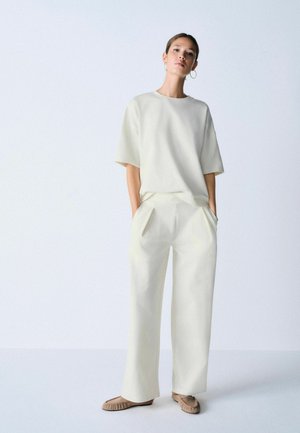 Žena stojaca s rukami v vreckách, oblečená v voľnom bielom tričku s krátkym rukávom, široké biele nohavice a béžové rovné topánky na jednofarebnom pozadí.