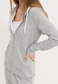 Sudadera gris con cremallera fabricada en material suave, con capucha con cordón blanco, bolsillos frontales y puños de canalé. Textura suave, diseño casual.
