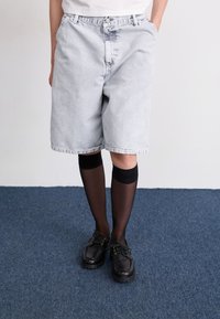 Heledad hall denim lühikesed püksid, lahtise lõikega, kuni põlveni ja peene pleekinud mustriga, koos mustade põlvikute ja massiivsete mustade kingadega.