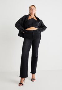 Mulher vestindo um top halter preto com pregas, jeans pretos de cintura alta, uma jaqueta preta com tachas colocada sobre os ombros, e sandálias pretas de salto alto.