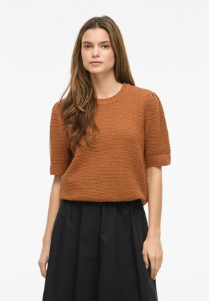 Woman with long brown hair wearing a textured rust-colored short-sleeve sweater and high-waisted black skirt standing against a white background.