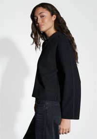 Maglione nero over size con spalle scese, finitura testurizzata e dettagli di cucitura sottili, indossato con jeans in denim scuro.