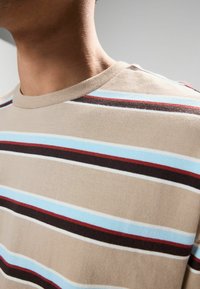 Beige t-shirt med korte ærmer og horisontale striber i lys blå, rød, brun og hvid. Blød tekstur og rund halsudskæring.