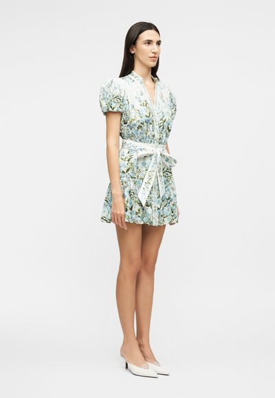 Woman standing in a short, floral dress with puff sleeves and a tied waist belt, wearing white pointed-toe low heels on a plain background.