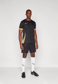 Mees seisab musta ja kullaga Puma jalgpallijerseys, mustades lühikestes pükstes, valgetes Puma sokkides ja mustades kingades, neutraalse tausta ees.