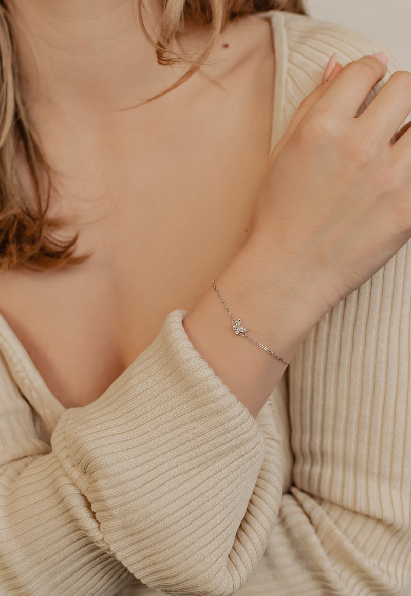 Silver bracelet with a small flower-shaped pendant, featuring a clear stone. The wrist is adorned with textured, ribbed beige fabric.