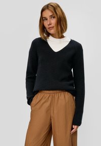 Zwarte gebreide trui met V-hals en lange mouwen, gecombineerd met tan broek met elastische taille. Gevoelige brei-details zijn zichtbaar.