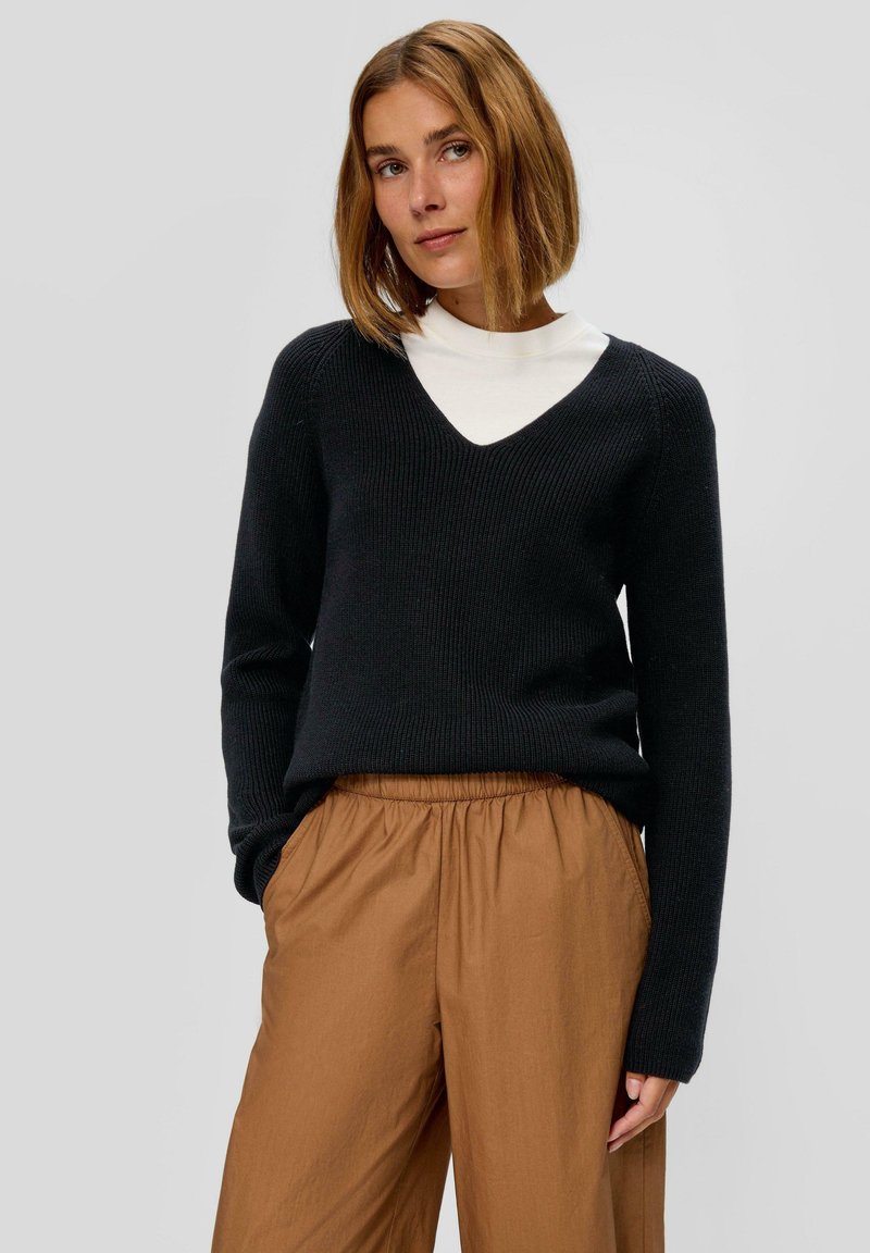 Zwarte gebreide trui met V-hals en lange mouwen, gecombineerd met tan broek met elastische taille. Gevoelige brei-details zijn zichtbaar.