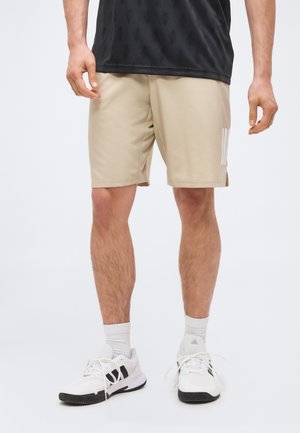 Mann trägt beige Sportshorts mit weißen Turnschuhen und weißen Socken, steht vor einem schlichten hellen Hintergrund.