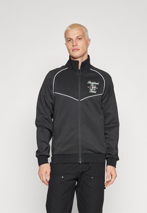 Must mustaga, millel on valged piping detailid, pehme kangas, kõrge krae ja broderitud logo vasakul rindkere piirkonnas.