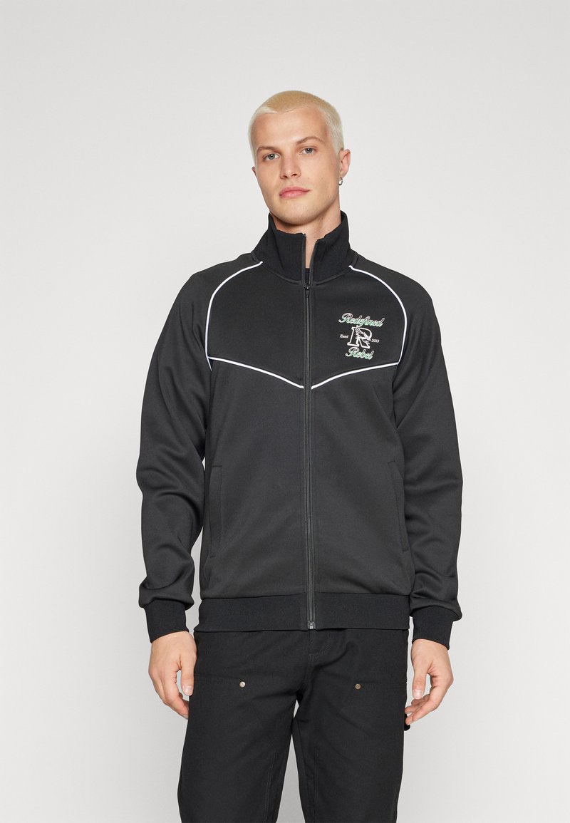 Must mustaga, millel on valged piping detailid, pehme kangas, kõrge krae ja broderitud logo vasakul rindkere piirkonnas.