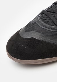 Zapatilla deportiva negra con parte superior de ante y malla, puntera redondeada, perforaciones para transpirabilidad y suela de goma con un acabado suave.