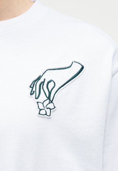 Χειροποίητη κέντηση ενός λεπτού χεριού που κρατάει ένα μικρό λουλούδι στο πάνω αριστερό μέρος ενός λευκού ενδύματος.