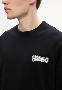 Felpa nera con collo a costine, caratterizzata da un logo ricamato bianco testurizzato "Hugo" sulla parte sinistra del petto.