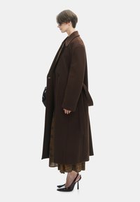 Bruine wollen overjas met een lang, oversized ontwerp; gecombineerd met een gebloemd jurkje en zwarte hoge hakken. De items hebben een minimalistische esthetiek.
