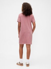 Woman with curly hair wearing a dusty pink short-sleeve dress and brown sneakers with white socks, facing away on a plain background.