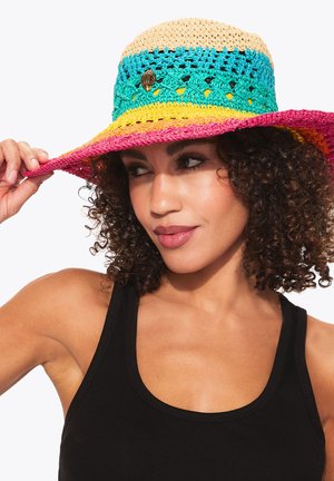 Donna con capelli ricci che indossa una canottiera nera e un cappello da sole all'uncinetto multicolore, tenendo la tesa con una mano.