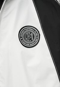 Μαύρο και άσπρο τμήμα αθλητικής ζακέτας που δείχνει κυκλικό έμβλημα του Jacker Football Club με τις λέξεις "Forever Hustling" και "2011".