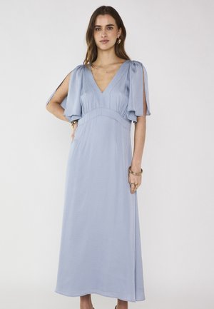 Femme portant une robe longue bleu clair à col en V, avec des manches fendues et volantées, des boucles d'oreilles anneaux dorées, ainsi que plusieurs bracelets et bagues en or.