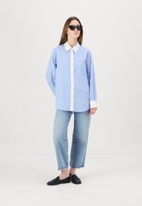 Camicia a righe bianco e blu con bottoni sul davanti, colletto e taschino sul petto. Abbinata a jeans azzurri chiari e scarpe slip-on nere.