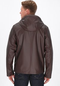 Giacca impermeabile marrone con cappuccio, realizzata in materiale liscio e flessibile. Presenta cuciture e un taglio dritto, indossata sopra jeans scuri.